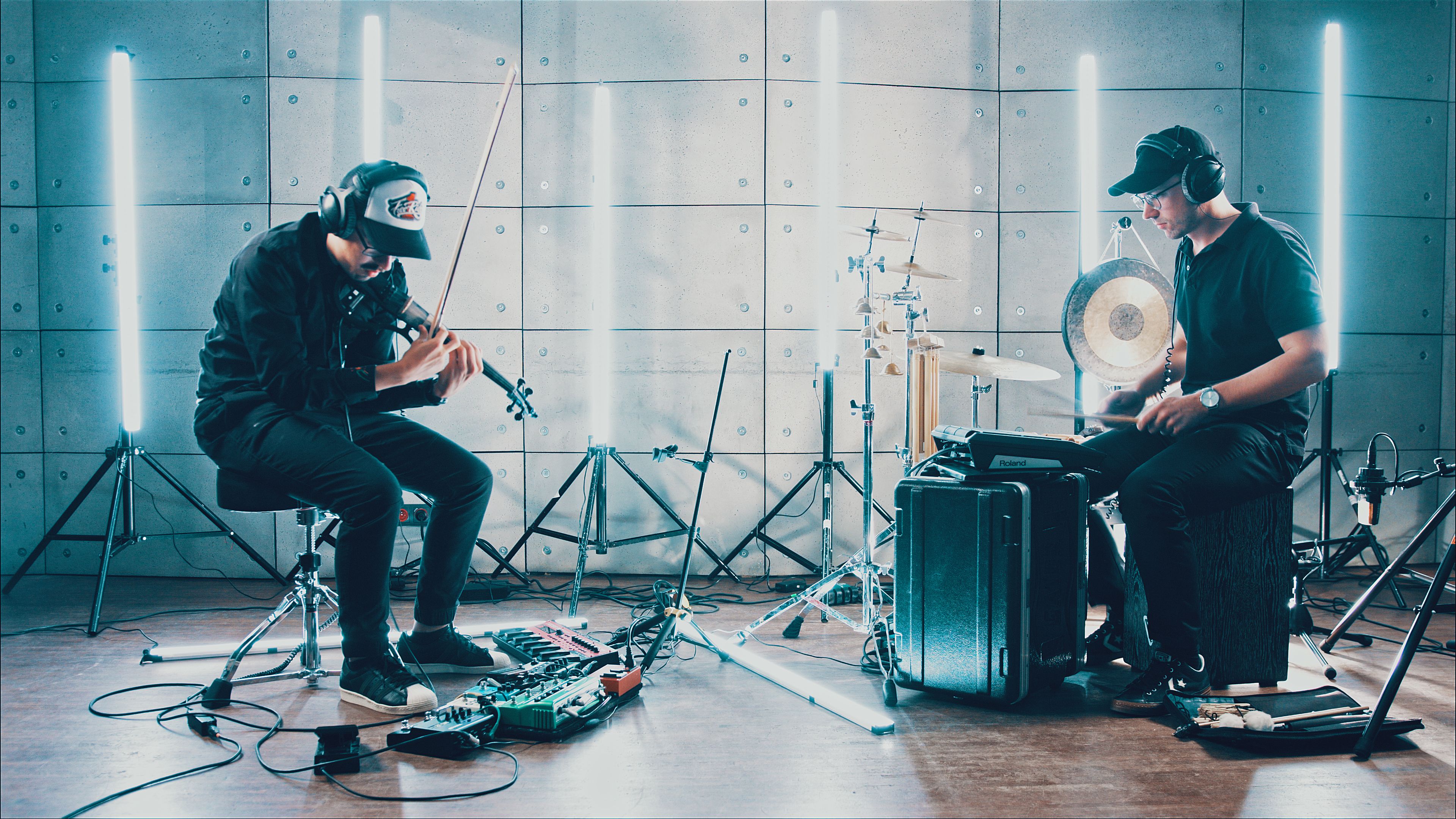 NØTNOIZ in the studio: a violinist with effects pedals and a percussionist with a gong and cymbals, lit by upright neon tubes